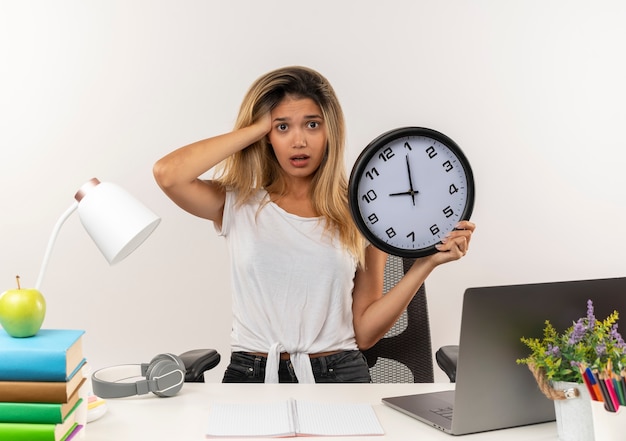 Impresionada joven estudiante bonita de pie detrás del escritorio con herramientas escolares poniendo la mano en la cabeza y sosteniendo el reloj aislado en blanco