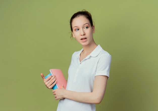 Impresionada joven estudiante bonita de pie en la vista de perfil sosteniendo el bloc de notas y el libro y mirando al lado aislado sobre fondo verde oliva con espacio de copia