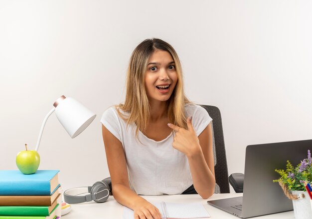 Impresionada joven estudiante bonita sentada en un escritorio con herramientas escolares apuntando a sí misma aislado en blanco