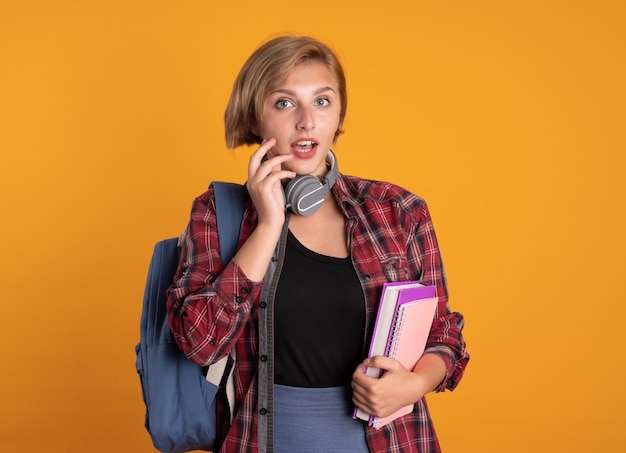 Impresionada joven estudiante eslava con auriculares con mochila sostiene libro y cuaderno