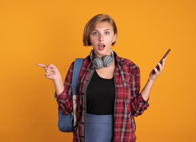 Impresionada joven estudiante eslava con auriculares con mochila sostiene el teléfono y apunta al lado