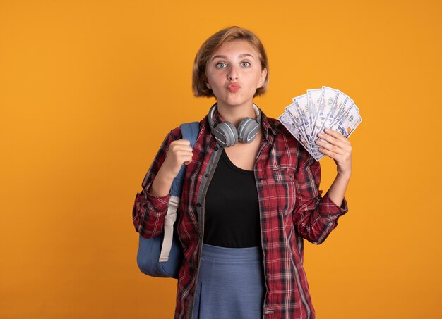 Impresionada joven estudiante eslava con auriculares con mochila tiene dinero