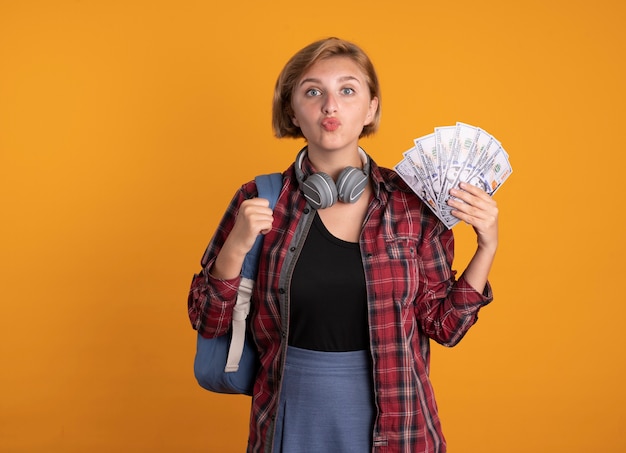 Impresionada joven estudiante eslava con auriculares con mochila tiene dinero