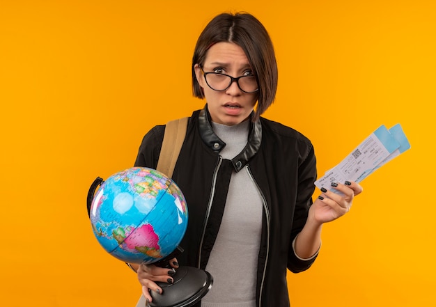 Foto gratuita impresionada joven estudiante con gafas y bolsa trasera con billetes de avión y globo aislado en naranja