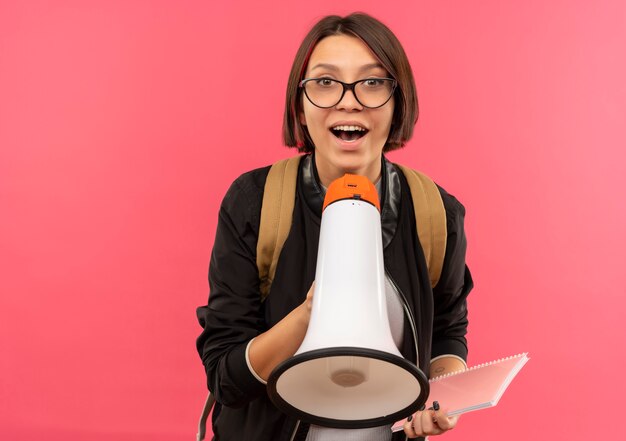 Impresionada joven estudiante con gafas y bolsa trasera con bloc de notas y altavoz aislado en rosa