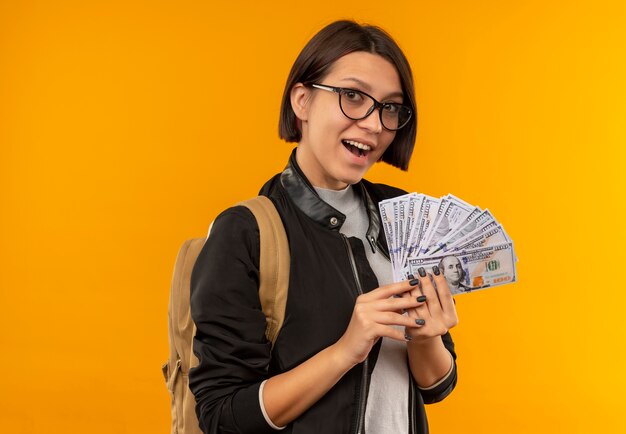 Impresionada joven estudiante con gafas y bolsa trasera con dinero aislado en naranja