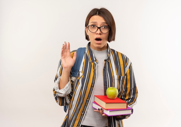 Impresionada joven estudiante con gafas y bolsa trasera con libros y manzana sobre ellos levantando la mano aislada en blanco