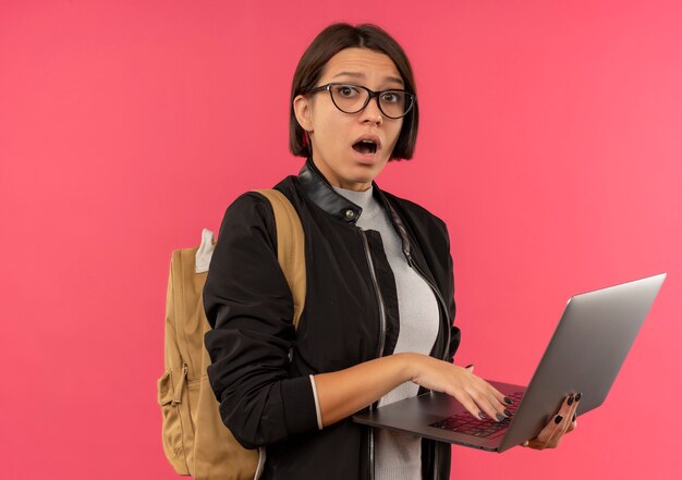 Impresionada joven estudiante con gafas y bolsa trasera con portátil aislado en rosa