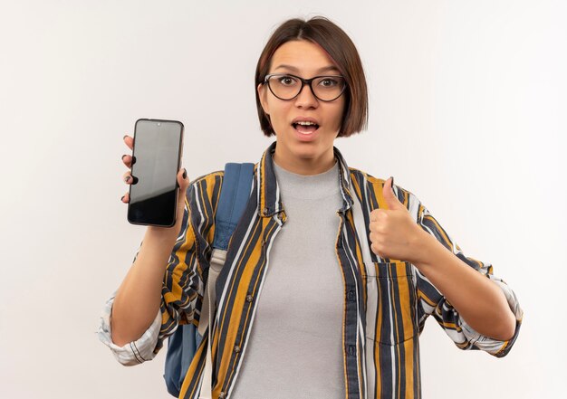 Impresionada joven estudiante con gafas y bolsa trasera que muestra el teléfono móvil y el pulgar hacia arriba aislado en blanco