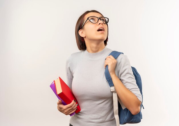 Impresionada joven estudiante con gafas y bolsa trasera sosteniendo libros mirando hacia arriba aislado en blanco