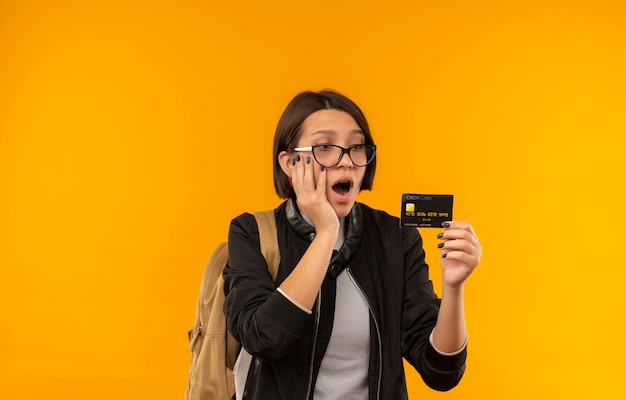 Impresionada joven estudiante con gafas y bolsa trasera sosteniendo y mirando la tarjeta de crédito con la mano en la cara aislada en naranja
