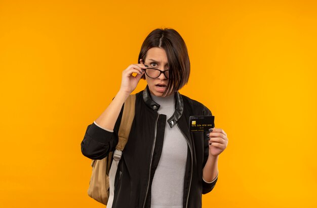 Impresionada joven estudiante con gafas y bolsa trasera con tarjeta de crédito poniendo la mano en vasos aislados en naranja