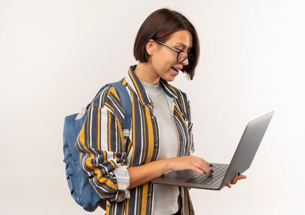 Impresionada joven estudiante con gafas y bolsa trasera usando laptop aislado en blanco