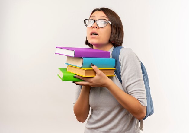 Impresionada joven estudiante con gafas y mochila sosteniendo libros aislados en blanco