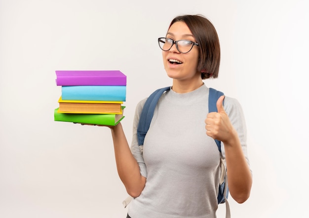 Impresionada joven estudiante con gafas y mochila sosteniendo libros mostrando el pulgar hacia arriba aislado en blanco