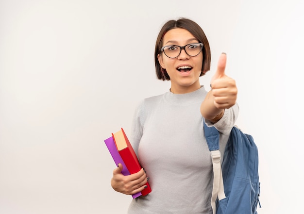 Foto gratuita impresionada joven estudiante con gafas y mochila sosteniendo libros mostrando el pulgar hacia arriba aislado en blanco