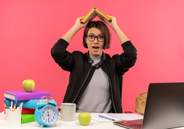 Impresionada joven estudiante con gafas sentado en el escritorio haciendo los deberes manteniendo el libro sobre la cabeza aislado en rosa