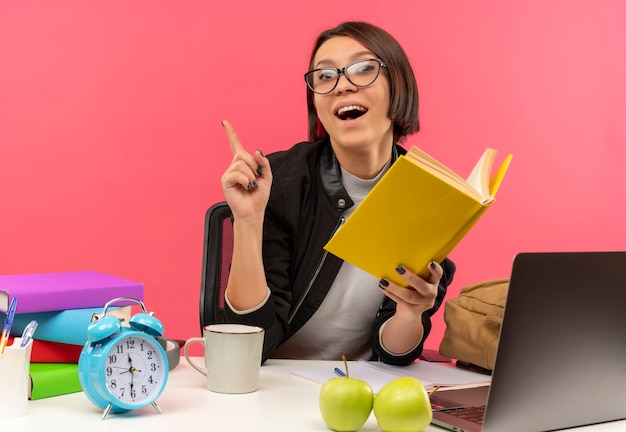 Impresionada joven estudiante con gafas sentado en el escritorio sosteniendo el libro haciendo los deberes levantando el dedo aislado en rosa