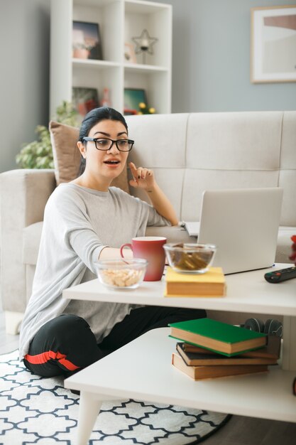 Impresionada joven con gafas utiliza portátil sentado en el suelo detrás de la mesa de café en la sala de estar