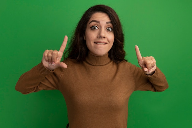 Impresionada joven hermosa apunta hacia arriba aislado en la pared verde