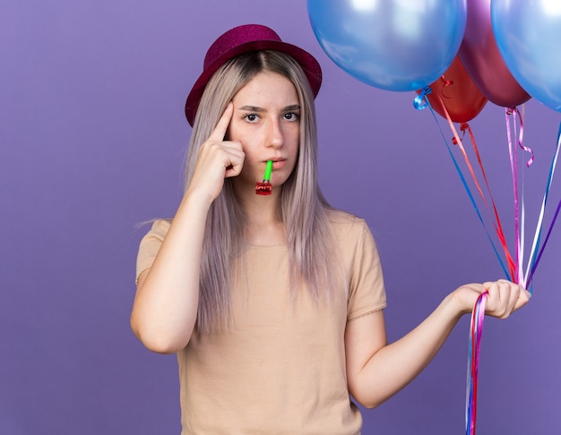 Impresionada joven hermosa con gorro de fiesta sosteniendo globos soplando silbato de fiesta poniendo el dedo en el templo aislado en la pared azul