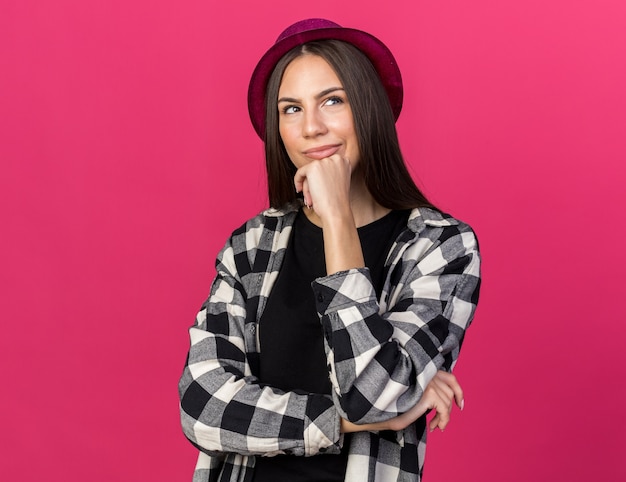 Impresionada joven hermosa mujer vistiendo gorro de fiesta poniendo la mano en la barbilla aislada en la pared rosa