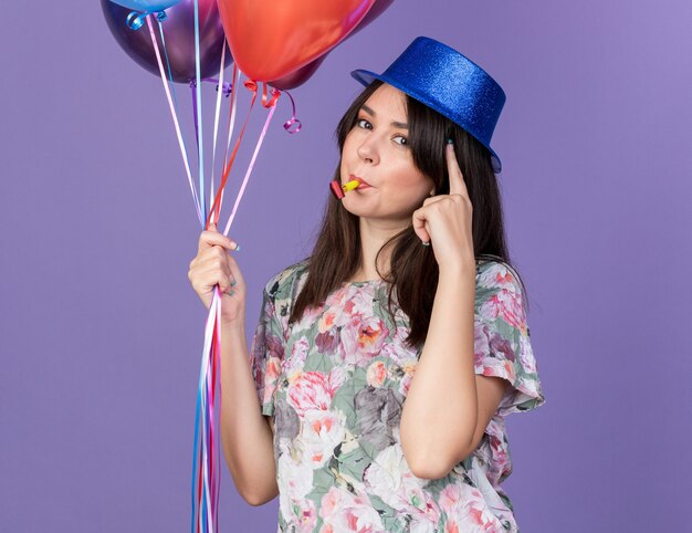 Impresionada joven hermosa mujer vistiendo gorro de fiesta sosteniendo globos soplando silbato de fiesta poniendo el dedo en el templo aislado en la pared azul