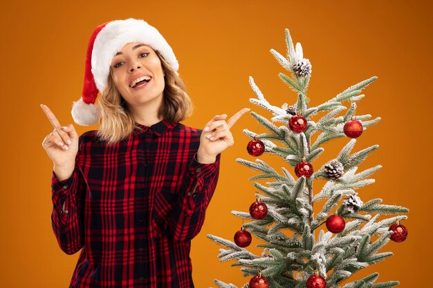 Impresionada joven hermosa de pie cerca del árbol de navidad vistiendo puntos de sombrero de navidad en diferentes lados aislado sobre fondo naranja