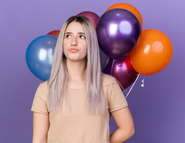 Impresionada joven hermosa de pie delante de globos aislados en la pared azul