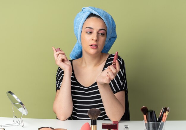 Impresionada joven hermosa se sienta a la mesa con herramientas de maquillaje envuelto el cabello en una toalla sosteniendo y mirando el lápiz labial aislado en la pared verde oliva