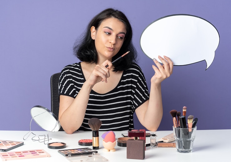 Foto gratuito impresionada joven hermosa se sienta a la mesa con herramientas de maquillaje sosteniendo bocadillo con pincel de maquillaje aislado en la pared azul