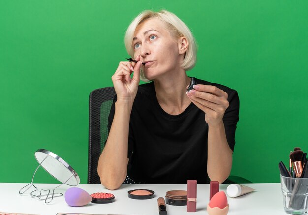 Impresionada joven hermosa se sienta a la mesa con herramientas de maquillaje sosteniendo rímel en la boca aislada en la pared verde