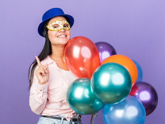 Impresionada joven hermosa con sombrero de fiesta y máscara de ojos de mascarada sosteniendo globos apunta hacia arriba aislado en la pared azul