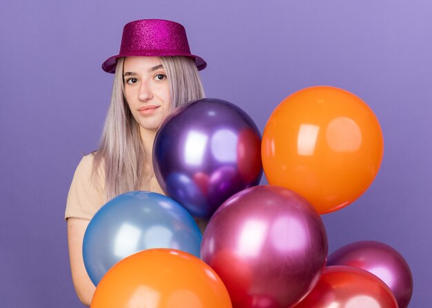 Impresionada joven hermosa con sombrero de fiesta de pie detrás de globos aislados en la pared azul