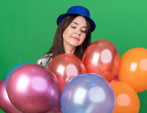 Impresionada joven hermosa con sombrero de fiesta de pie detrás de globos aislados en la pared verde