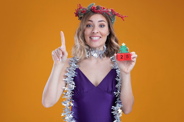 Foto gratuita impresionada joven hermosa vestida de púrpura vestido y corona con guirnalda en el cuello sosteniendo puntos de juguete de navidad en arriba aislado sobre fondo marrón