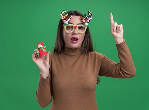 Impresionada joven hermosa vestida de suéter marrón y gafas de Navidad sosteniendo puntos de juguete de Navidad en arriba aislado sobre fondo verde