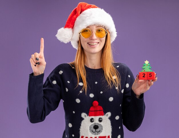 Impresionada joven hermosa vestida con un suéter de Navidad y sombrero con gafas sosteniendo puntos de juguete de Navidad en arriba aislado sobre fondo púrpura