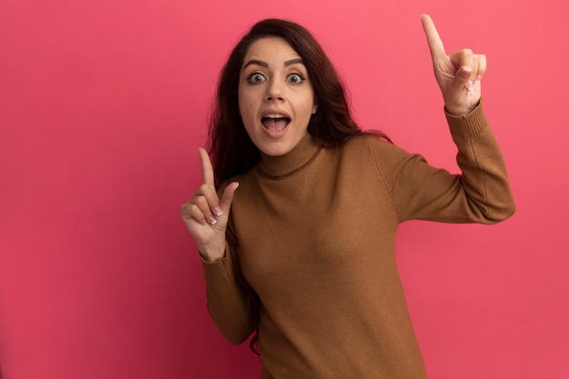 Impresionada joven hermosa vistiendo suéter de cuello alto apunta hacia arriba aislado en la pared rosa