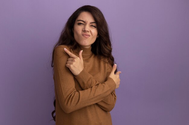 Impresionada joven hermosa vistiendo suéter de cuello alto marrón puntos en diferentes lados aislados en la pared púrpura con espacio de copia