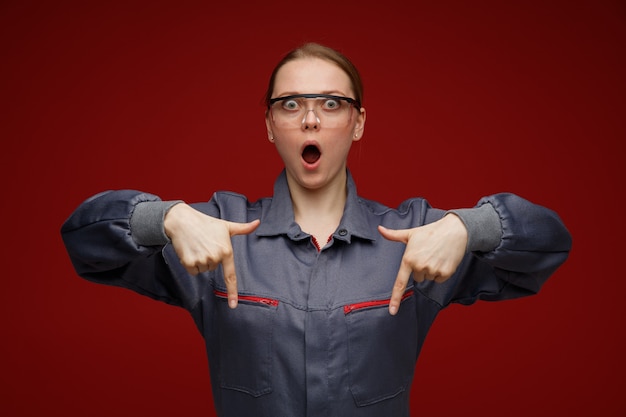 Impresionada joven ingeniera rubia vestida con uniforme y gafas de seguridad apuntando hacia abajo