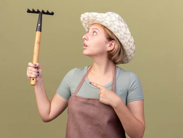 Impresionada joven jardinera eslava con sombrero de jardinería sosteniendo y apuntando al rastrillo aislado en la pared verde oliva con espacio de copia