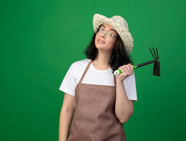 Impresionada joven jardinera morena con gafas ópticas y uniforme con sombrero de jardinería tiene rastrillo de azada aislado en la pared verde