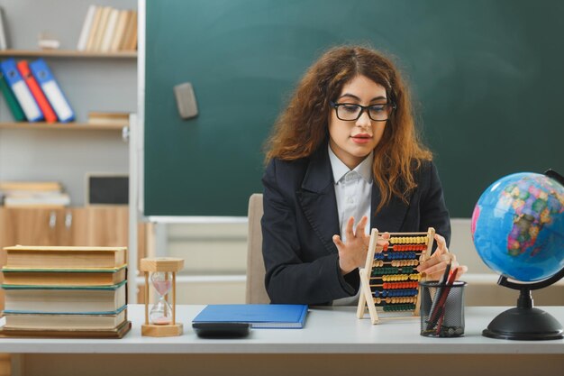 impresionada joven maestra con gafas sosteniendo y mirando abacus sentado en el escritorio con herramientas escolares en el aula