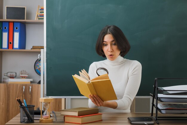 impresionada joven maestra mirando un libro con lupa sentada en el escritorio con herramientas escolares en el aula