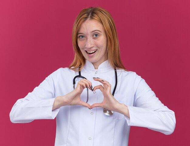 Impresionada joven médico de jengibre vistiendo bata médica y estetoscopio mirando al frente haciendo signo de corazón aislado en la pared carmesí