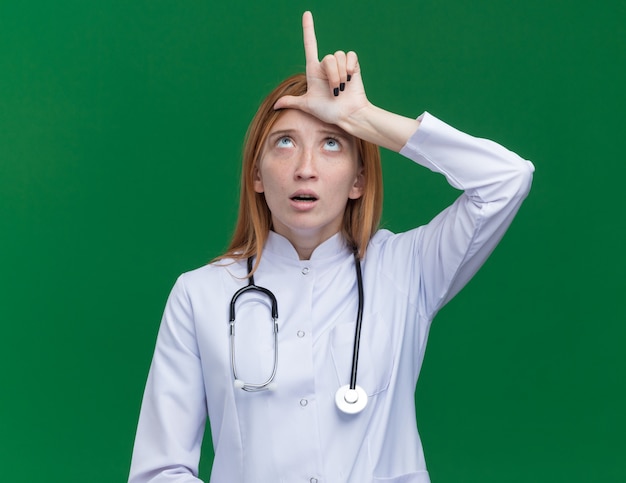 Impresionada joven médico de jengibre vistiendo bata médica y un estetoscopio mirando hacia arriba haciendo gesto de perdedor en la frente