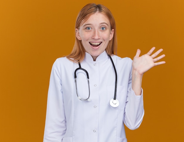 Impresionada joven médico de jengibre vistiendo bata médica y un estetoscopio mostrando la mano vacía aislada en la pared naranja