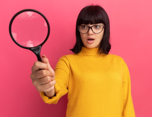 Impresionada joven morena caucásica en gafas ópticas sostiene y mira lupa aislada en pared rosa con espacio de copia