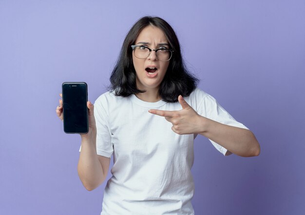 Impresionada joven mujer bonita con gafas mostrando y apuntando al teléfono móvil